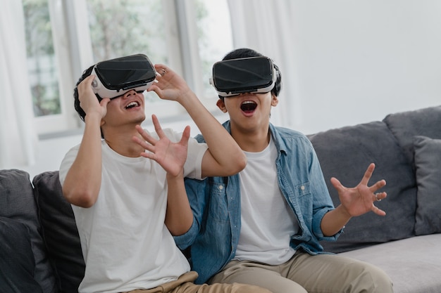 Молодая пара азиатских геев, использующих технологии, смешные дома, парень-любитель Азии lgbtq +, чувствующий себя счастливым в виртуальной реальности, играющий в игры вместе, лежа на диване в гостиной дома.