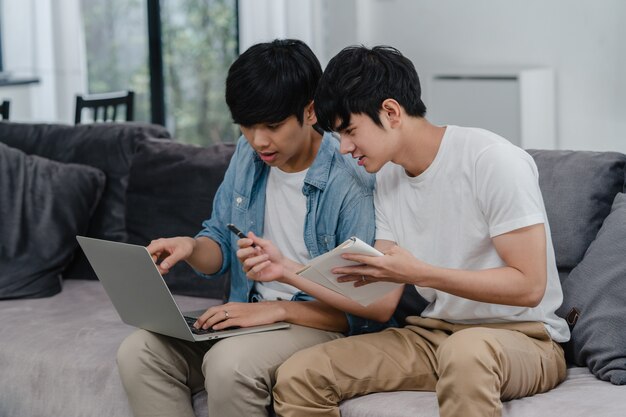 現代の家でラップトップを働く若いアジアゲイカップル。アジアLGBTQ +の男性は、コンピューターを使用して、家のリビングルームでソファに横たわっている間インターネットで彼らの財政を一緒に分析して楽しくリラックスします。