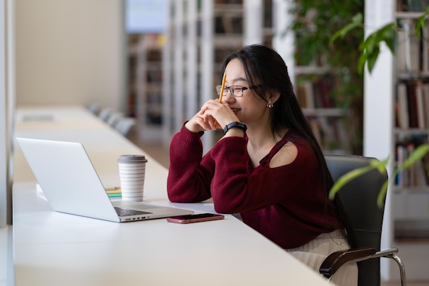 図書館​で​ノート​パソコン​の​勉強​を​見て​笑う​若い​アジア​の​女の子​は​、​オンラインウェビナー​や​講義​を​見て​楽しんでいます