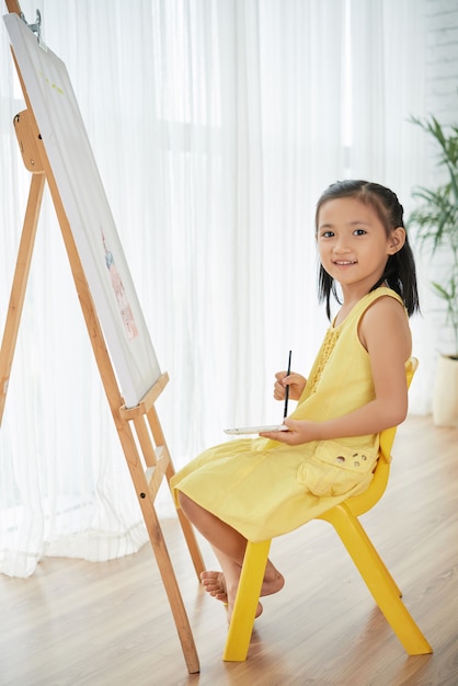 Young Asian girl posing at home in front of easel, with painting brush and watercolors