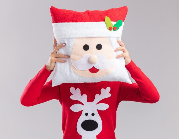 白い背景で隔離のクリスマス枕とセーターで覆われた顔でクリスマス帽子をかぶって若いアジアの女の子