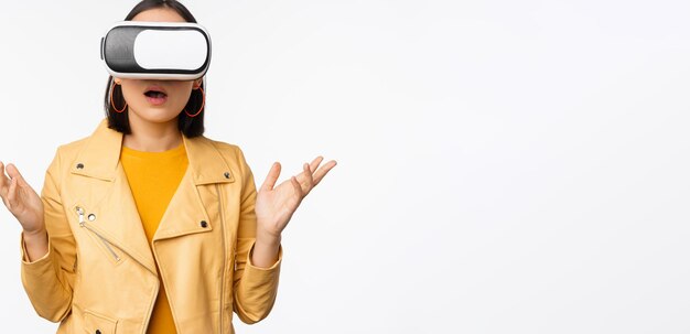 Young asian girl wearing vr glasses looking amazed using virtual reality headset posing against whit