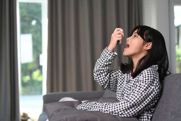 Young asian girl with asthma using asthma inhaler for preventing attacks on sofa.