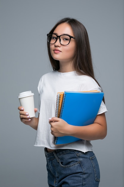 Молодая азиатская девушка с тетрадью и кофе для того чтобы пойти в руки стоя изолирована против серой предпосылки