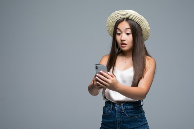 Молодая азиатская девушка с телефоном пользы соломенной шляпы на серой предпосылке
