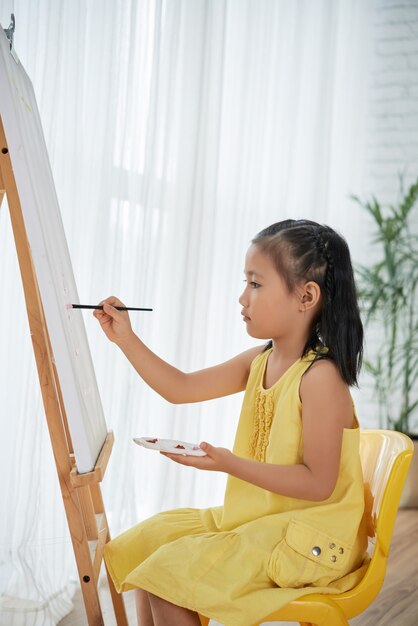 Young Asian girl in yellow dress sitting in front of easel at home and painting