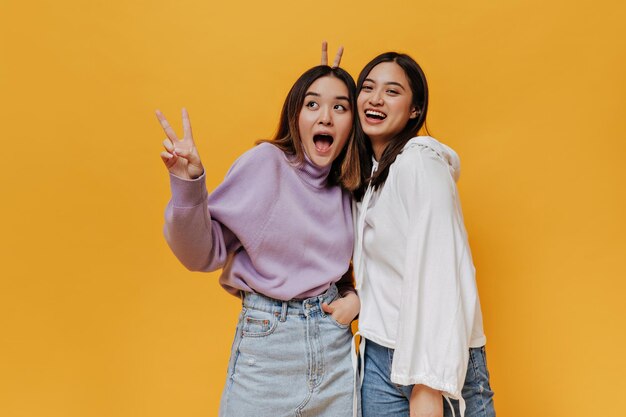 Young Asian girls laugh and pose on orange background Pretty brunette woman in purple sweater shows peace sign Goodhumored girl in white hoodie gives bunny ears to friend