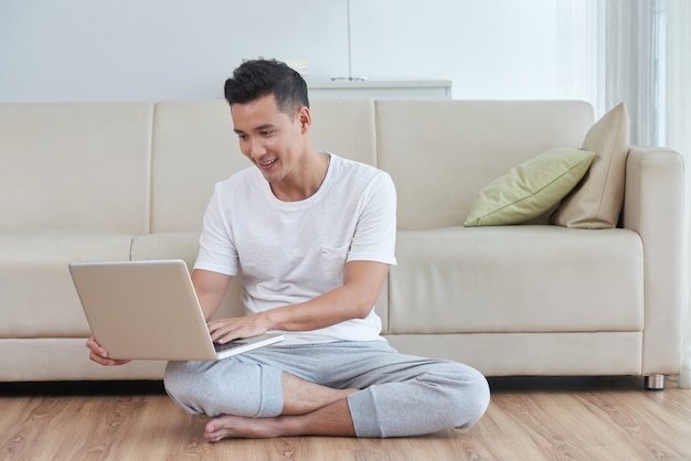 Young Asian guy using his laptop on the floor of the living room next to the beige sofa