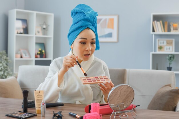Young asian happy smiling woman with towel on her head sitting at the dressing table at home interior applying eyeshadows looking at mirror doing morning makeup routine