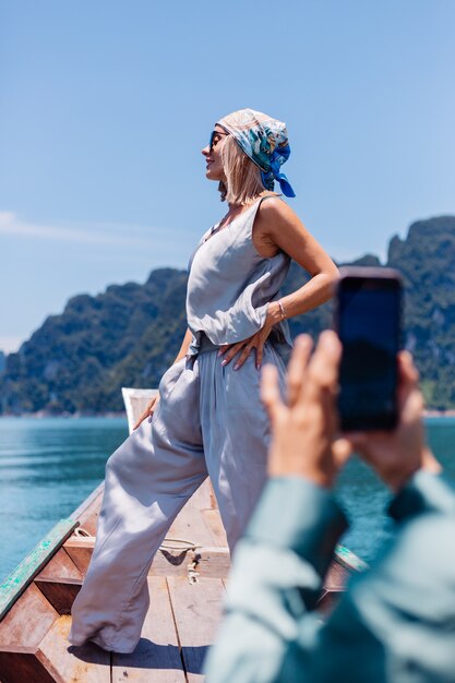 Молодой азиатский счастливый турист блоггера женщины в шелковом костюме и шарфе и солнечных очках на каникулах путешествует вокруг Таиланда на азиатской лодке, национальном парке Khao Sok.