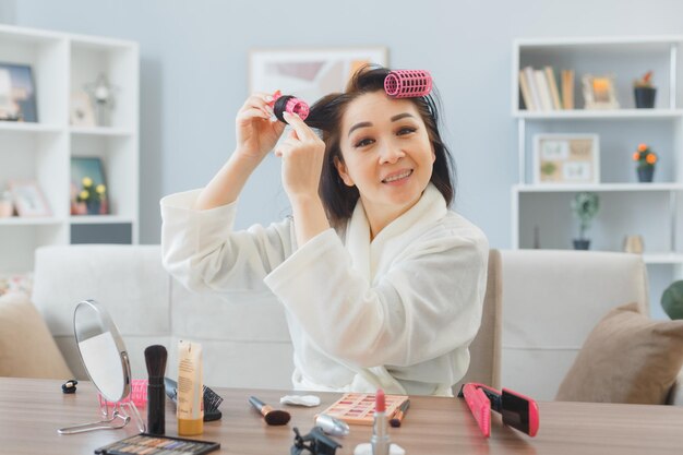 긴 검은 머리를 가진 젊은 아시아 행복한 여성이 집 내부 화장대에 앉아 아침 화장을 하는 머리에 롤러를 바르고 있다