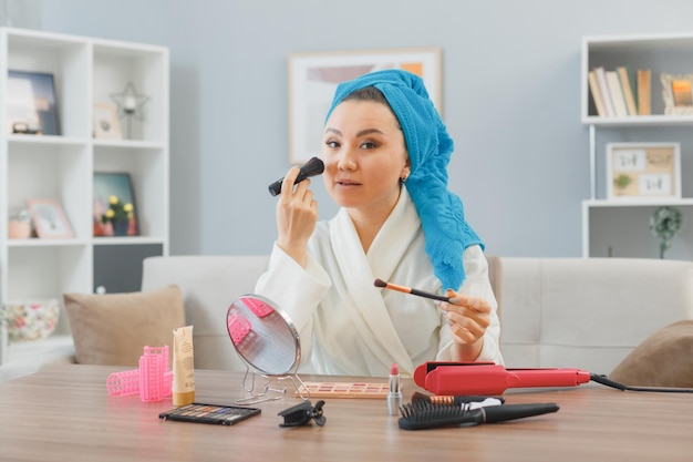 Young asian happy woman with towel on her head sitting at the dressing table at home applying eyeshadows and blusher doing morning makeup routine beauty and facial cosmetics concept