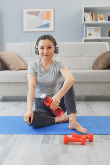 헤드폰을 끼고 집에서 실내 운동을 하는 젊은 아시아 여성, 요가 매트에 앉아 있는 거실에서 물 한 병과 아령