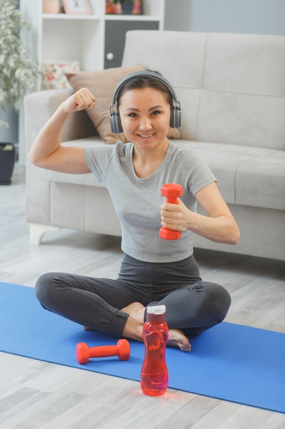 彼女の強さと上腕二頭筋を示すヨガマットの上に座っているリビングルームで水のボトルとダンベルで自宅で屋内で運動しているヘッドフォンを持つ若いアジアの健康な女性