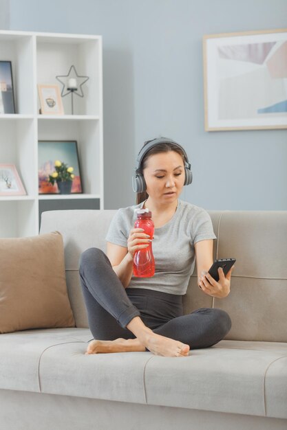 Young asian healthy woman with headphones sitting on a couch with bottle of water at home interior tired and overworked after workout using smartphone