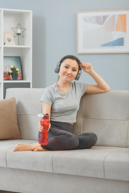 Young asian healthy woman with headphones sitting on a couch with bottle of water at home relaxing after workout happy and positive smiling