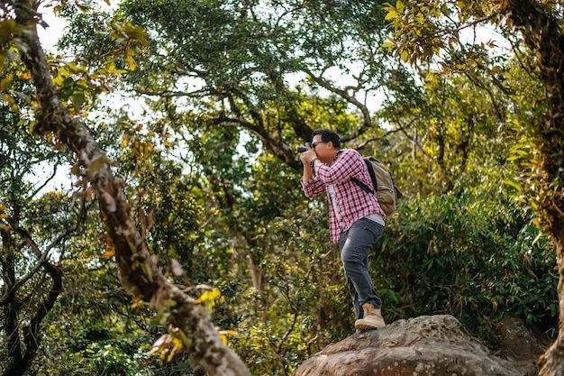 若いアジアのハイカーの男とバックパックは、森のコピースペースで写真を撮るためにカメラを使用しています