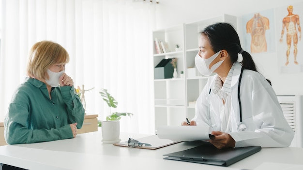 병원 사무실에서 여자 환자와 결과 또는 증상을 논의하는 클립 보드를 사용하여 보호 마스크를 착용하는 젊은 아시아 여자 의사.