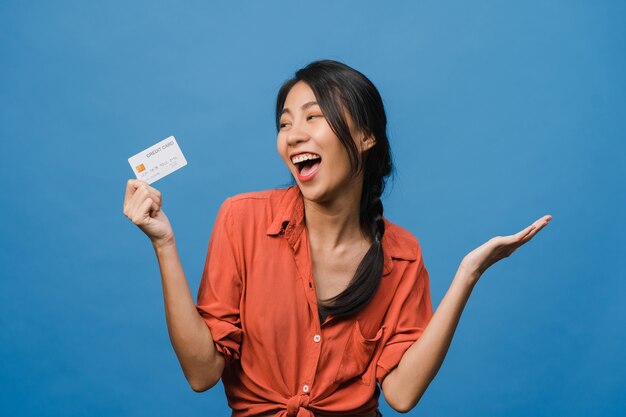 若いアジアの女性は、前向きな表情でクレジットカードを表示し、広く笑顔で、幸せを感じるカジュアルな服を着て、青い壁に孤立して立っています。表情のコンセプト。