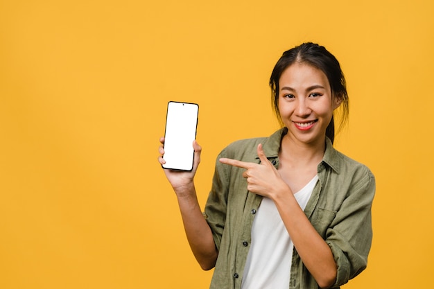 Young Asian lady show empty smartphone screen with positive expression, smiles broadly, dressed in casual clothing feeling happiness on yellow wall. Mobile phone with white screen in female hand.