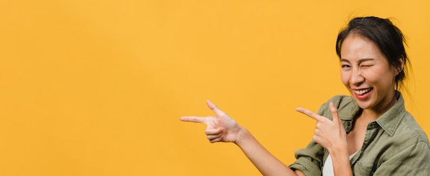 Free Photo young asian lady smiling with cheerful expression, shows something amazing at blank space in casual cloth  isolated over yellow wall. panoramic banner with copy space.