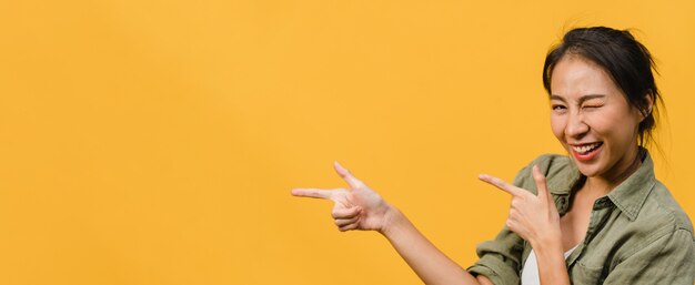 Young Asian lady smiling with cheerful expression, shows something amazing at blank space in casual cloth  isolated over yellow wall. Panoramic banner with copy space.