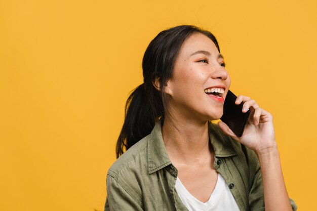 若いアジア人女性は、前向きな表情で電話で話し、広く笑顔で、幸せを感じるカジュアルな服を着て、黄色い壁に孤立して立っています。幸せな愛らしい嬉しい女性は成功を喜んでいます。