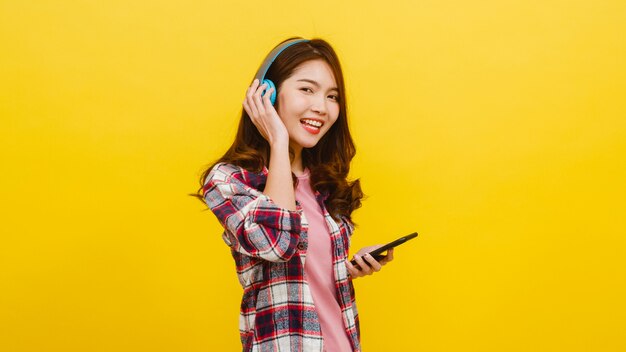 Young Asian lady wearing wireless headphones listening to music from smartphone with cheerful expression in casual clothing and looking at camera over yellow wall. Facial expression concept.