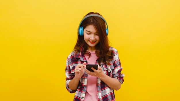 カジュアルな服装で陽気な表情でスマートフォンから音楽を聴くと黄色の壁越しにカメラを見てワイヤレスヘッドフォンを着ている若いアジア女性。顔の表情のコンセプトです。