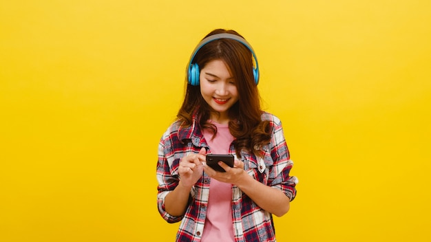 Young Asian lady wearing wireless headphones listening to music from smartphone with cheerful expression in casual clothing and looking at camera over yellow wall. Facial expression concept.