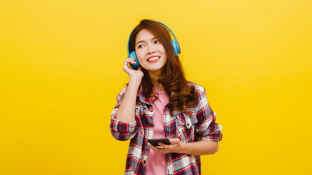 Young Asian lady wearing wireless headphones listening to music from smartphone with cheerful expression in casual clothing and looking at camera over yellow wall. Facial expression concept.