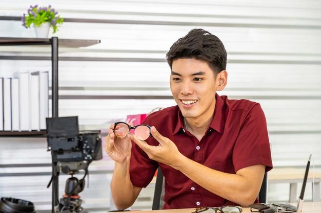 若いアジア人男性のオンライン商人ブロガーがカメラを使ってライブビデオで眼鏡をレビューして販売するソーシャルメディアインフルエンサーとオンラインショッピングのコンセプト