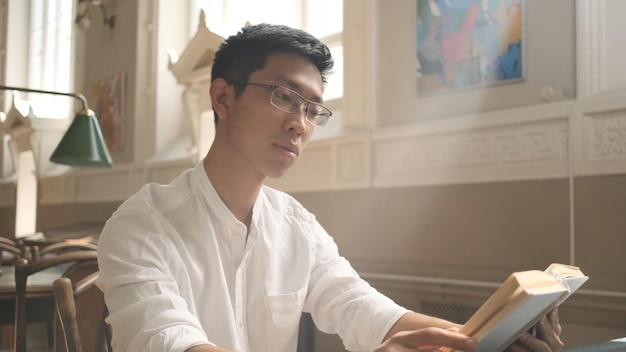 Молодой азиатский студент внимательно читает книгу в библиотеке университета Привлекательный парень готовится к экзаменам в кампусе колледжа
