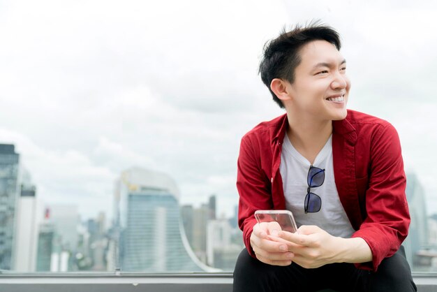 若いアジア人男性カジュアルリラックス赤いシャツ手持ちガラススマートフォン都市のダウンタウンのビジネス技術のアイデアの概念を背景に