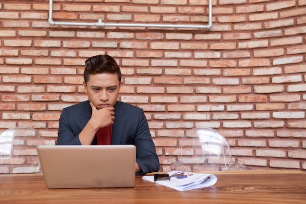 Young Asian man looking at laptop screen and rubbing his chin