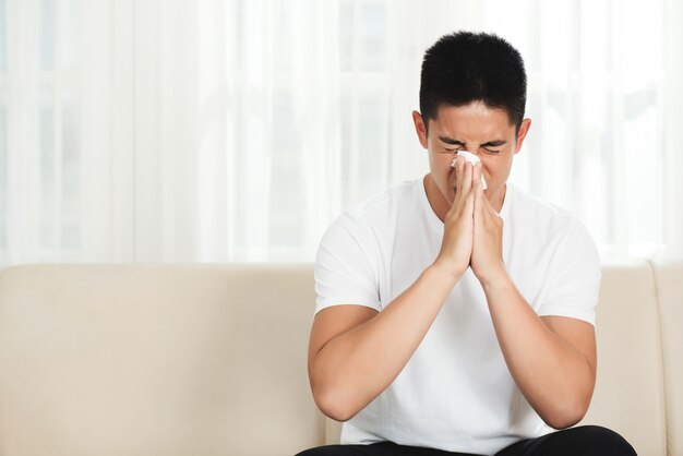 Young Asian man sitting on couch at home and blowing nose with tissue