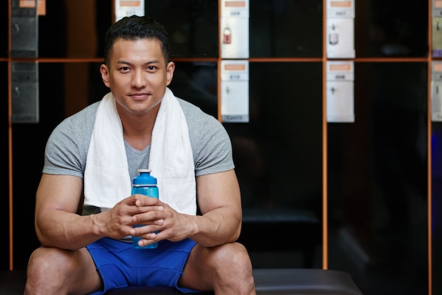 Young Asian man sitting in locker room in gym with water bottle and towel