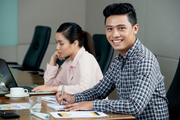 Young Asian man sitting at meeting table in office and smiling, and woman working on laptop