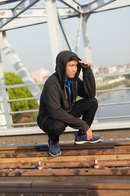 Free photo young asian man with hooded jacket crouched on a railway looking far away with hand on forehead