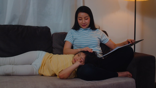 Young Asian mother read fairy tales to her daughter at home. Happy Chinese family relax with teen girl who sleep while listening to tales lying on bed in bedroom at modern house at night .