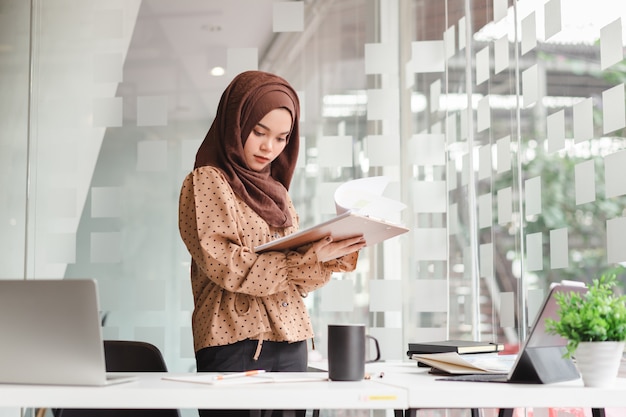 茶色​の​ヒジャーブカジュアル​な​服装​で​ビジネス​を​議論し​、​創造的​な​カフェ​に​立っている​間​笑顔​で​若い​アジア​の​イスラム​教徒​の​ビジネス​女性​。