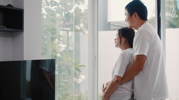 젊은 아시아 임신 몇 포옹과 그들의 아이와 이야기 배꼽 잡고. 엄마와 아빠가 집에서 거실에서 창문 가까이 아기, 임신을 돌보는 동안 평화로운 미소 행복 느낌.