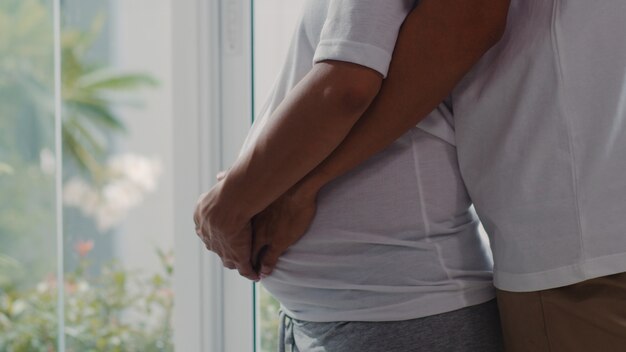 Young Asian Pregnant couple hug and holding belly talking with their child. Mom and Dad feeling happy smiling peaceful while take care baby, pregnancy near window in living room at home .