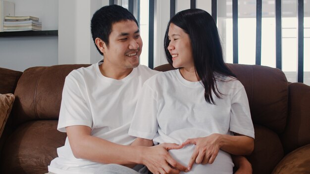 Young Asian Pregnant couple making heart sign holding belly. Mom and Dad feeling happy smiling peaceful while take care baby, pregnancy lying on sofa in living room at home .