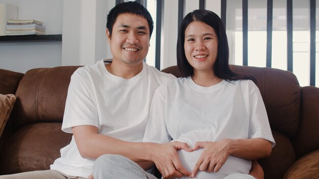 腹を保持している心のサインを作る若いアジア妊娠カップル。お母さんとお父さんは、赤ちゃんの世話をしながら平和な幸せな笑顔を感じて、妊娠は自宅のリビングルームのソファに横たわっています。