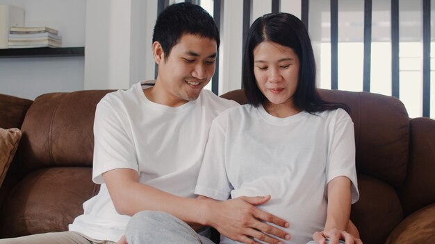Young Asian Pregnant couple man touch his wife belly talking with his child. Mom and Dad feeling happy smiling peaceful while take care baby, pregnancy lying on sofa in living room at home .