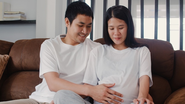 젊은 아시아 임신 몇 사람이 그의 아이와 이야기하는 그의 아내 배꼽을 터치합니다. 엄마와 아빠가 아기를 돌보는 동안 평화로운 미소 행복을 느끼고, 집에서 거실에 소파에 누워있는 임신.