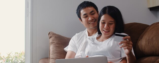 Молодая азиатская беременная пара онлайн покупки дома. Мама и папа чувствуют себя счастливыми, используя технологию ноутбука и кредитную карту, покупая детское изделие, лежа на диване в гостиной дома.