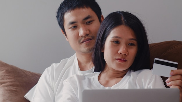 Young Asian Pregnant couple online shopping at home. Mom and Dad feeling happy using laptop technology and credit card buying baby product while lying on sofa in living room at home .