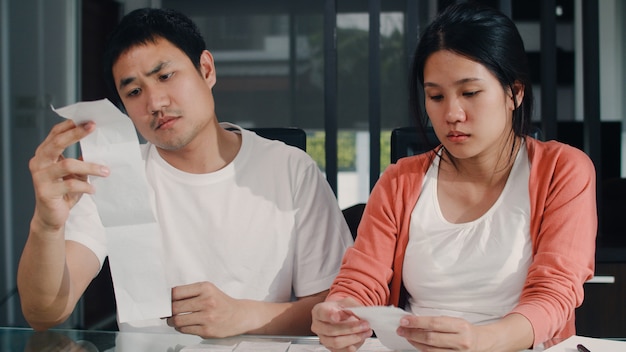 Young Asian Pregnant couple records of income and expenses at home. Dad worried, serious, stress while record budget, tax, financial document working in living room at home .